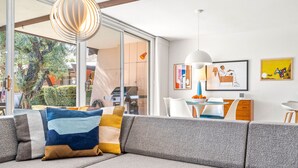 A modern living room with a grey sectional sofa, a dining area with a white table and chairs, and a large window with a view of greenery.