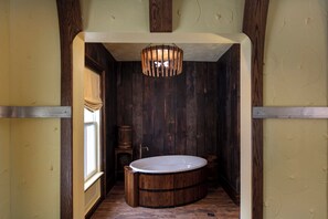 A wooden bathtub in a narrow room with a wooden ceiling and walls, a window on the left, and a hanging light fixture.