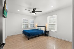 Une chambre à coucher comprenant un lit, un ventilateur de plafond, deux tables de chevet avec des lampes et deux fenêtres équipées de stores.