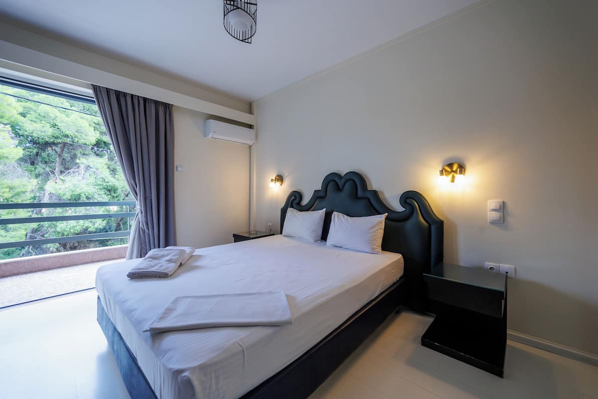 Un lit bien fait, avec du linge de lit blanc, une table de chevet avec une lampe, et une vue sur la verdure par la fenêtre.