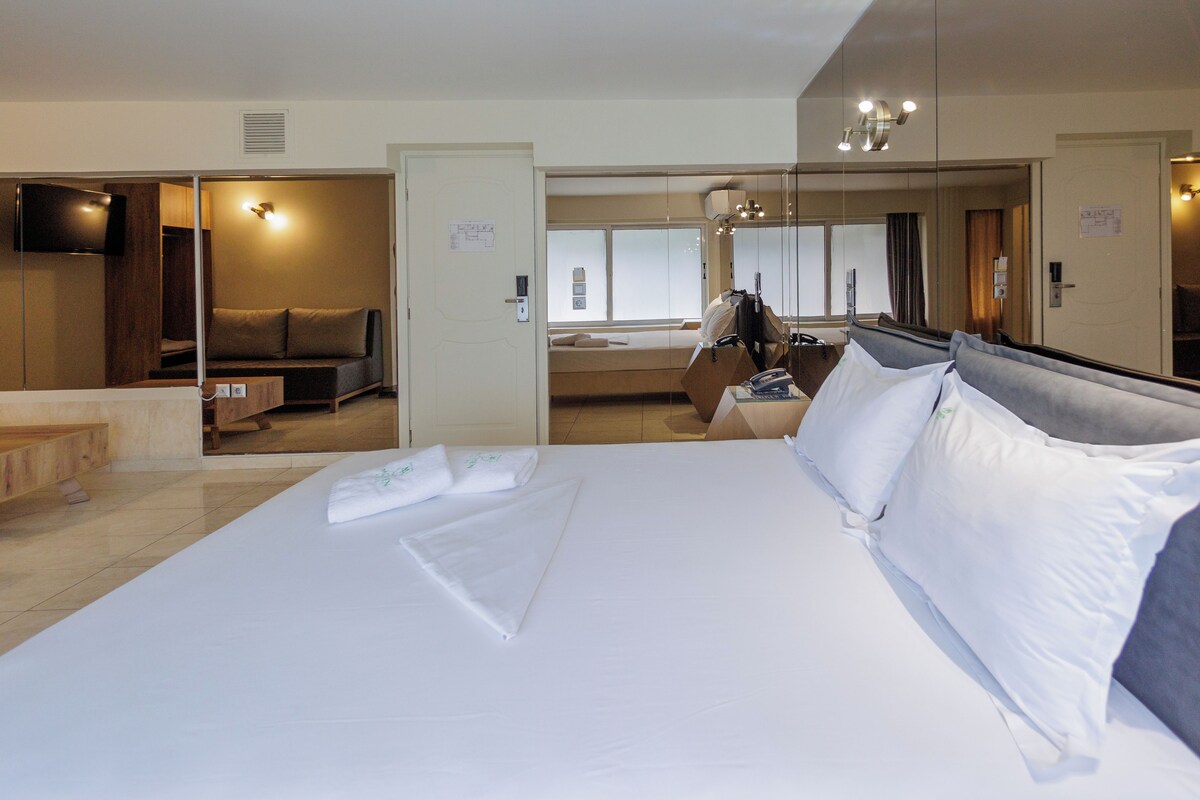 A hotel room with a large bed, a seating area, a TV, and a mirror.
