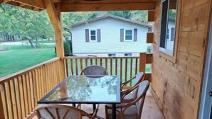 Une terrasse en bois couverte, avec une table en verre et des chaises en osier, donnant sur une cour verdoyante et une maison.