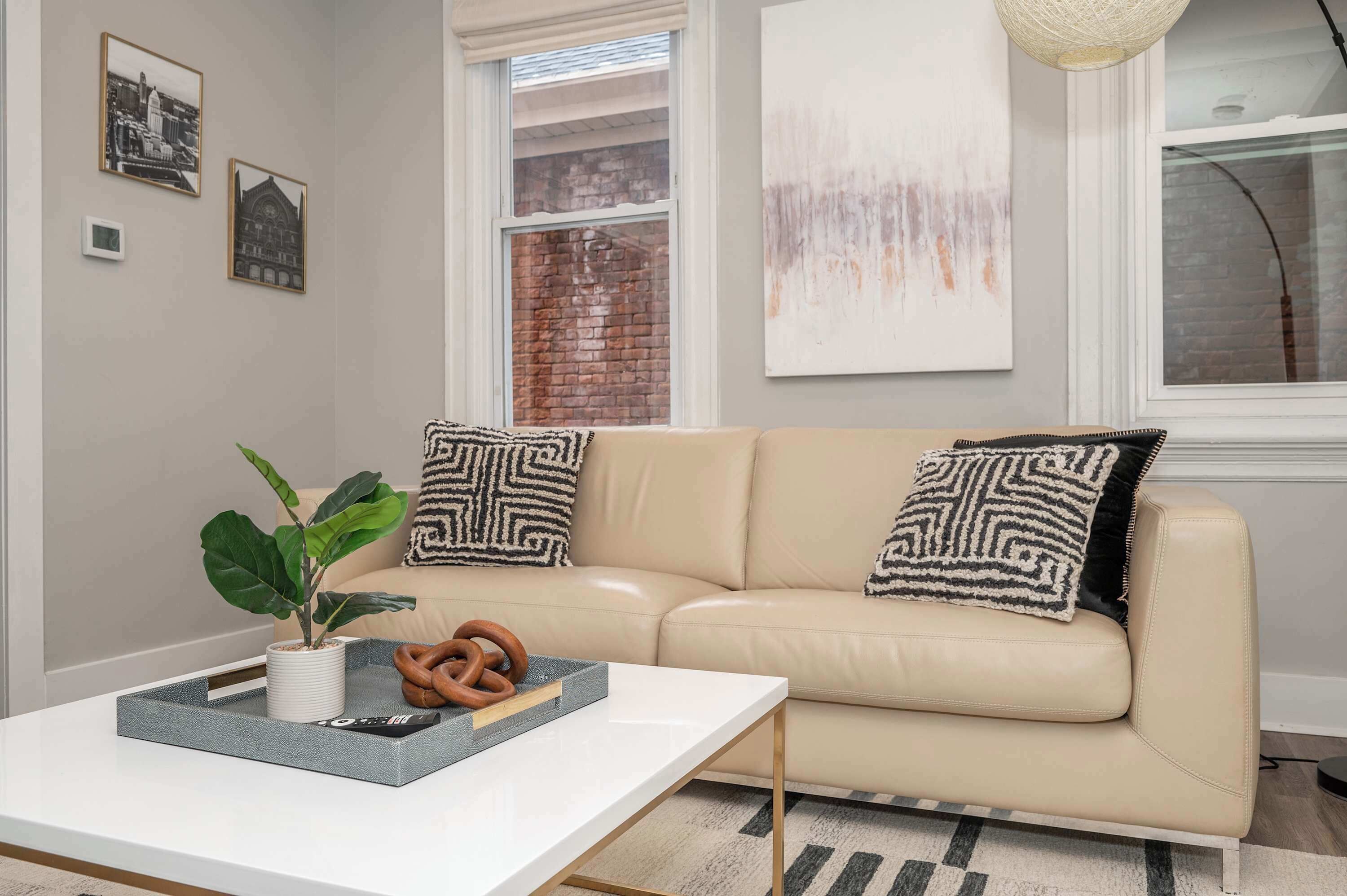 A modern living room with a beige leather sofa, two patterned throw pillows, a white coffee table with a tray holding a plant, a remote control, and a book.