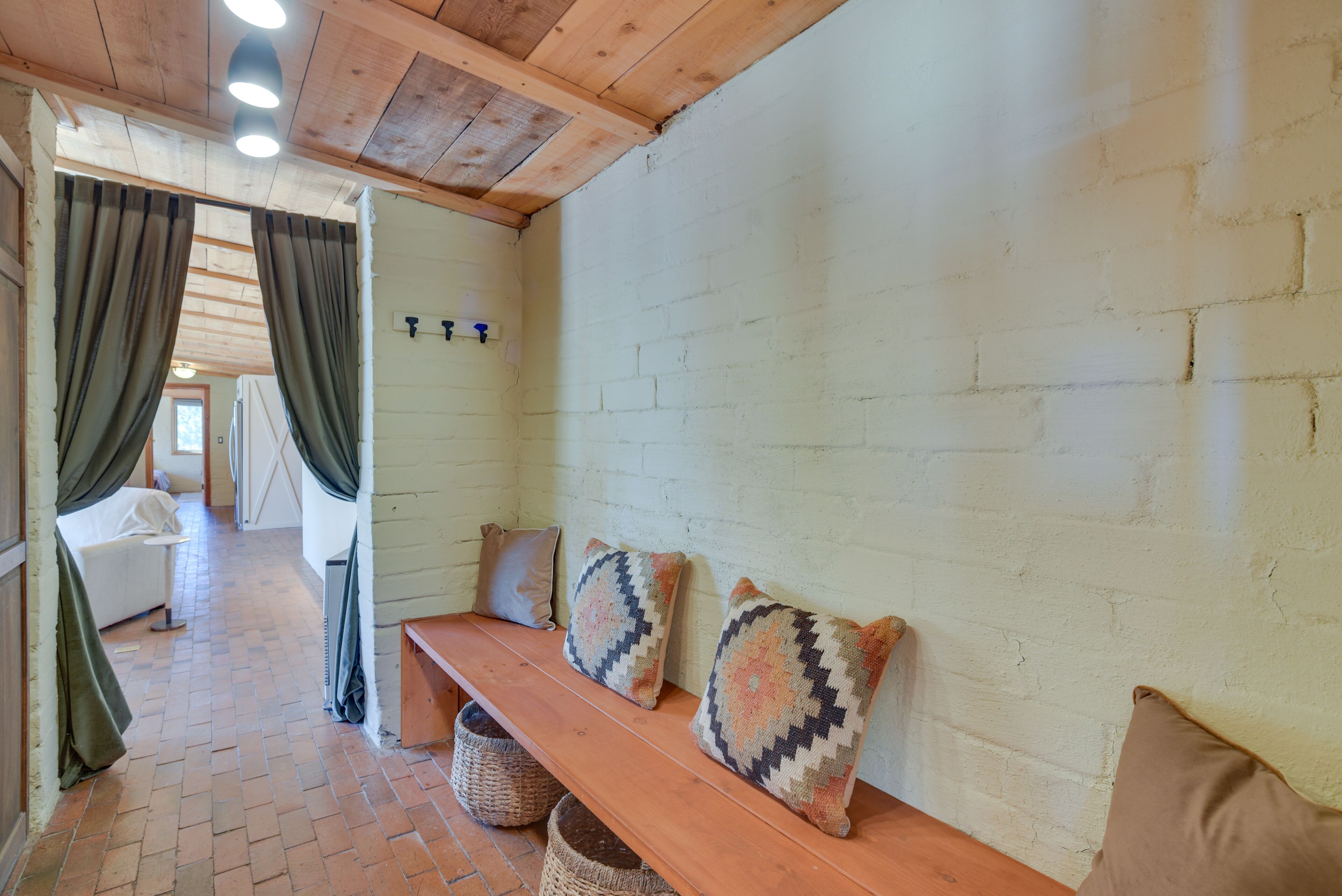 Una habitación acogedora con un banco de madera, dos almohadones estampados, una pared de ladrillo blanco y una ventana con cortinas.