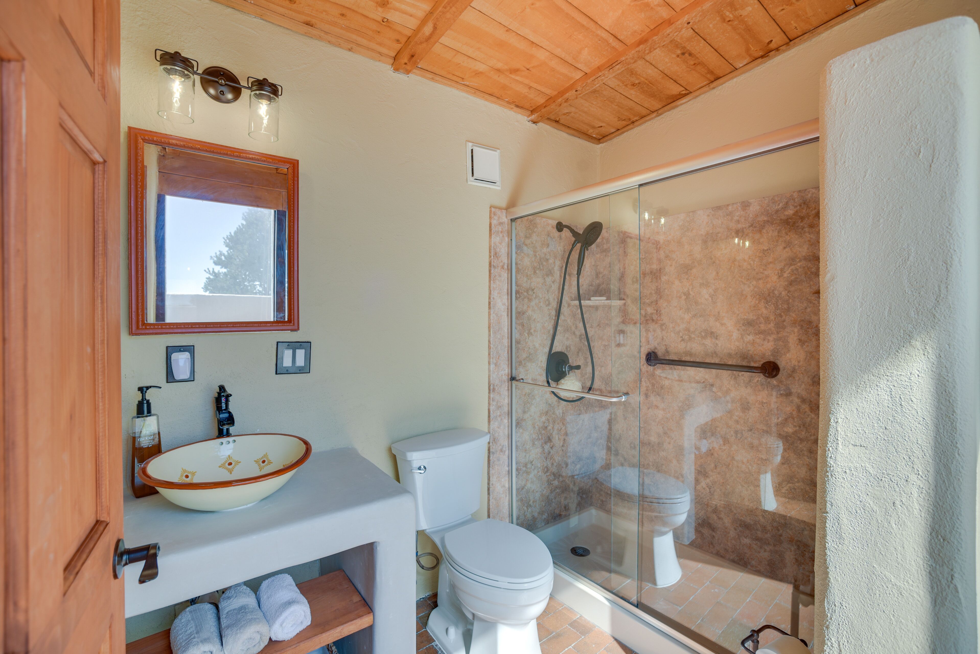 Un baño con techo de madera, ducha con cerramiento de vidrio, lavabo blanco con cuenca y un inodoro.