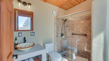 Un baño con techo de madera, ducha con cerramiento de vidrio, lavabo blanco con cuenca y un inodoro.