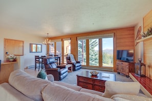Een ruime woonkamer met een grote bank, een eethoek met stoelen, een televisie en uitzicht op de omgeving.