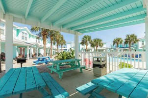 Un espace extérieur abrité, doté de bancs turquoise et de tables de pique-nique, d’une piscine et d’une clôture blanche.