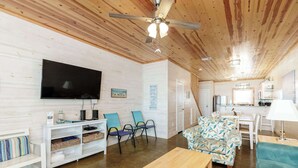 A cozy living room with a flat-screen TV, wooden ceiling, and a dining area with a kitchen in the background. - Resort Pool & Pet Friendly Sleeps 9 (Port Aransas)