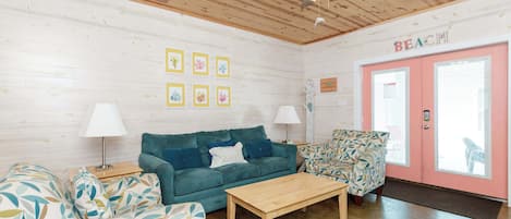 A cozy room with a wooden ceiling, a teal sofa, a patterned armchair, a wooden coffee table, and a pink door labeled 'BEACH'.