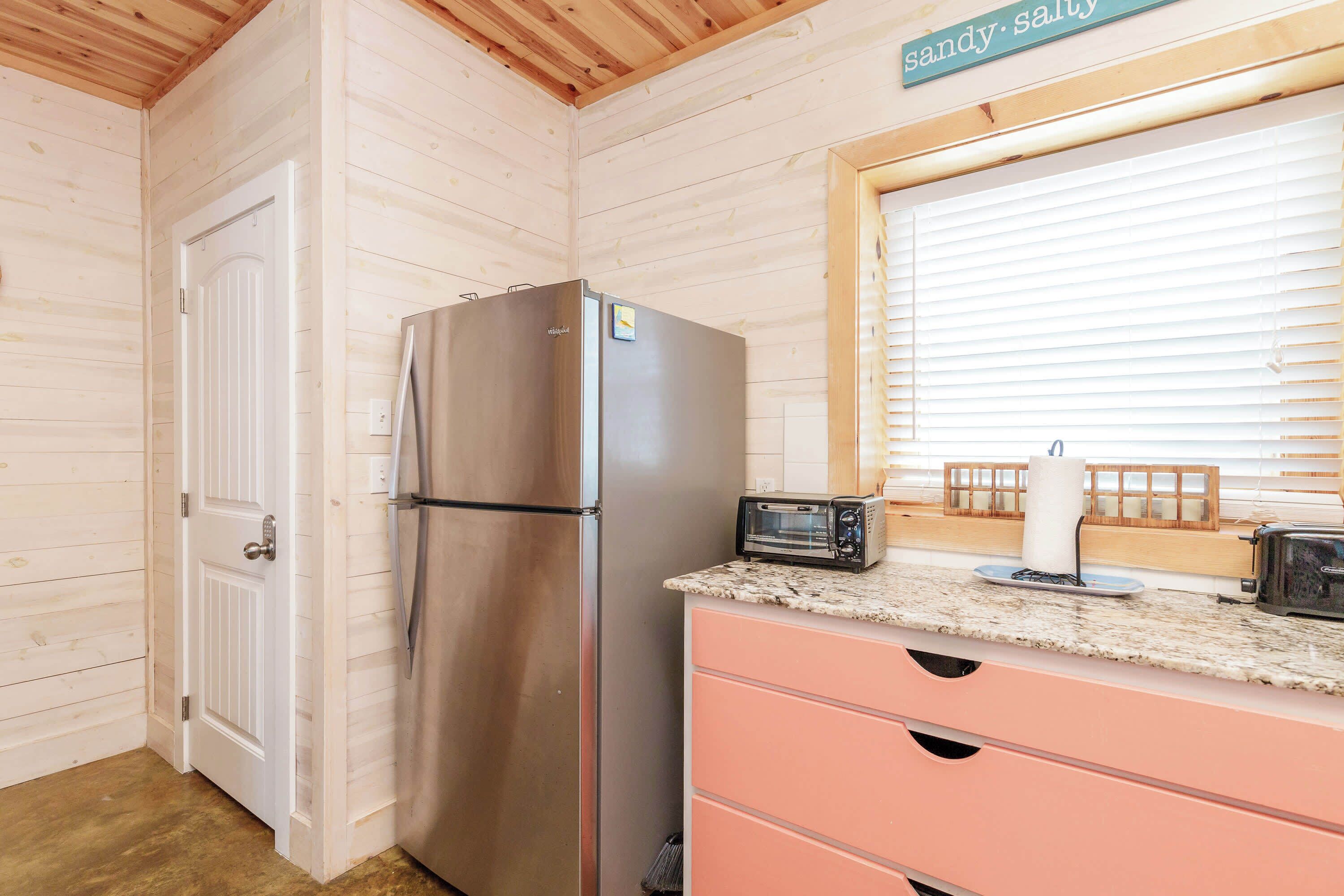 A compact kitchen with a stainless steel refrigerator, a toaster, and a wooden-framed window.