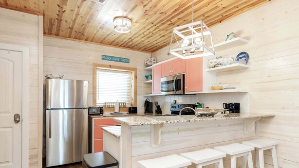 A kitchen with wooden walls, a stainless steel refrigerator, a microwave, and a sink. There are three white stools and a window with blinds. - Resort Pool & Pet Friendly Sleeps 9 (Port Aransas)