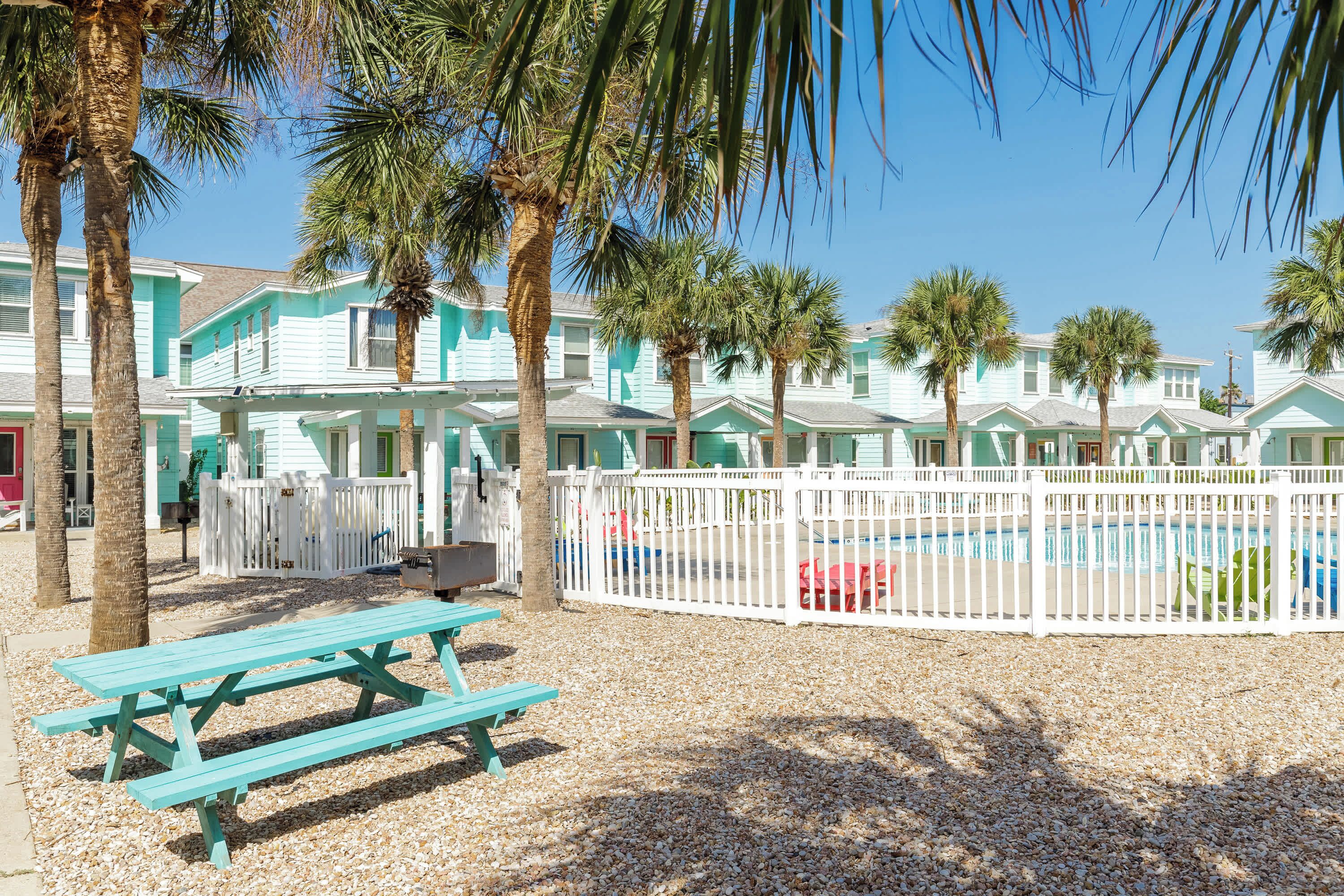 A row of turquoise beach houses with a pool, palm trees, and a picnic table.