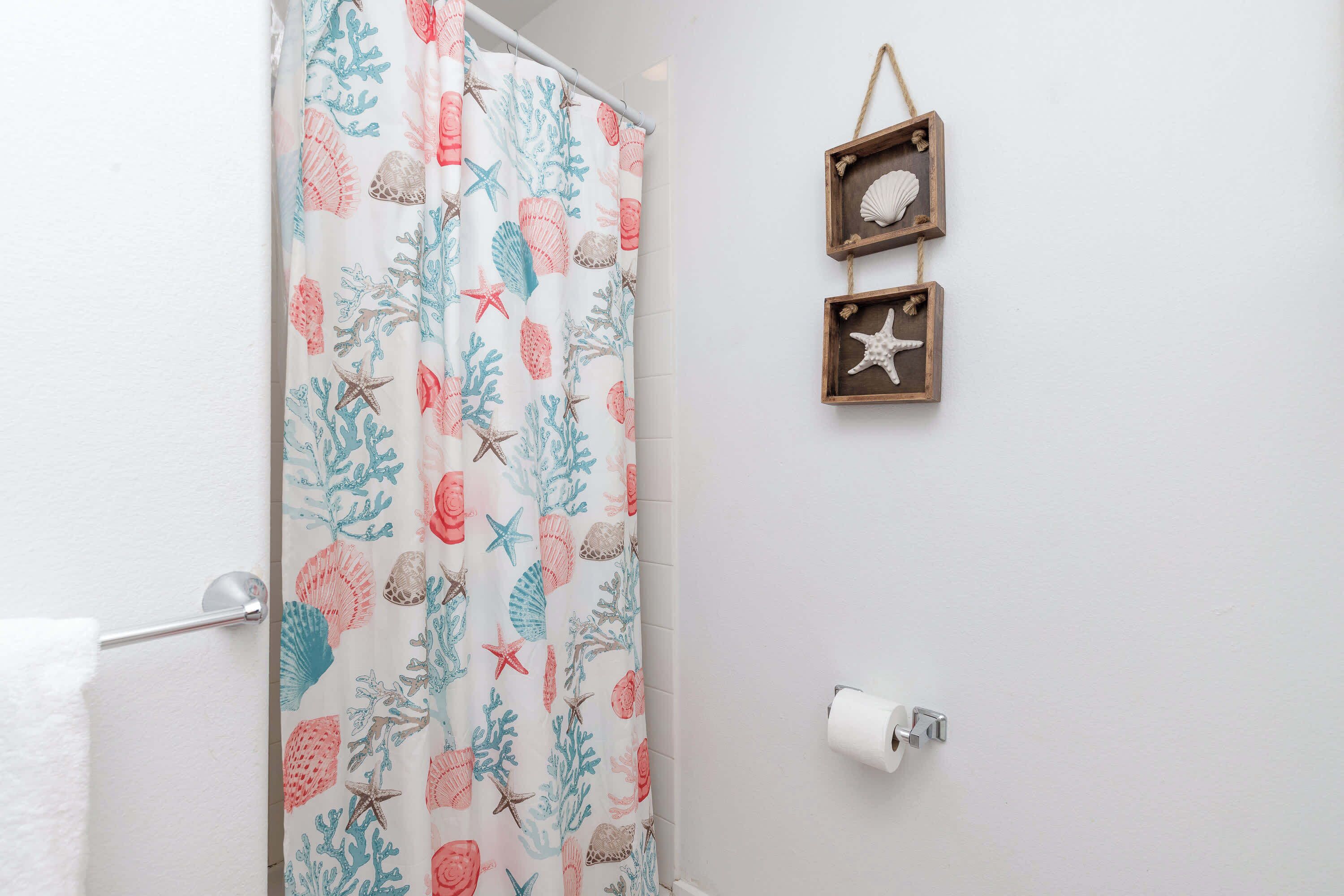 A bathroom with a shower curtain featuring a sea shell and starfish pattern, a toilet paper holder, and two wooden frames with shells on the wall.