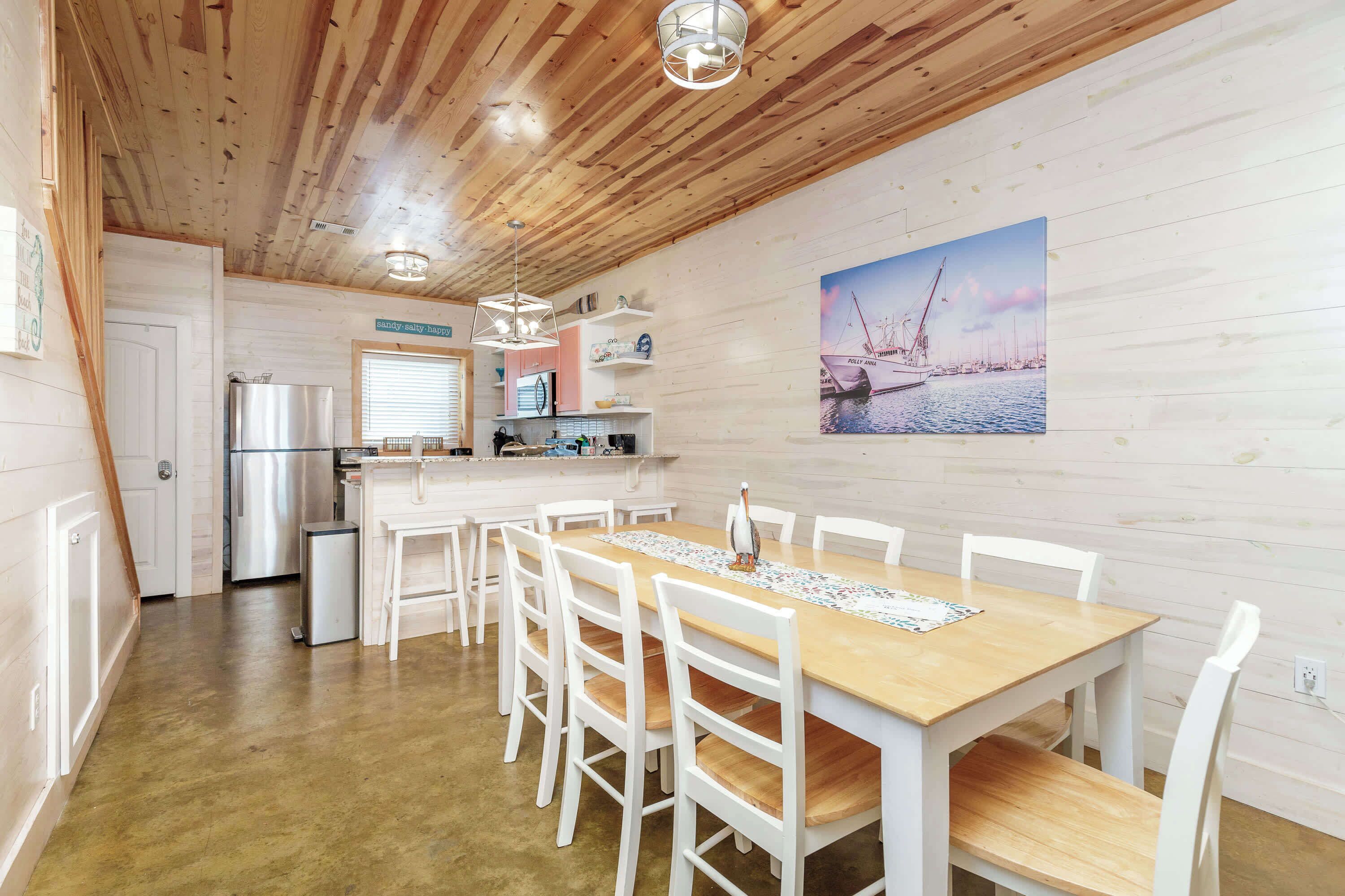 A dining area with a wooden table and white chairs, a kitchen with a refrigerator and a sink, and a wall-mounted painting of a harbor.