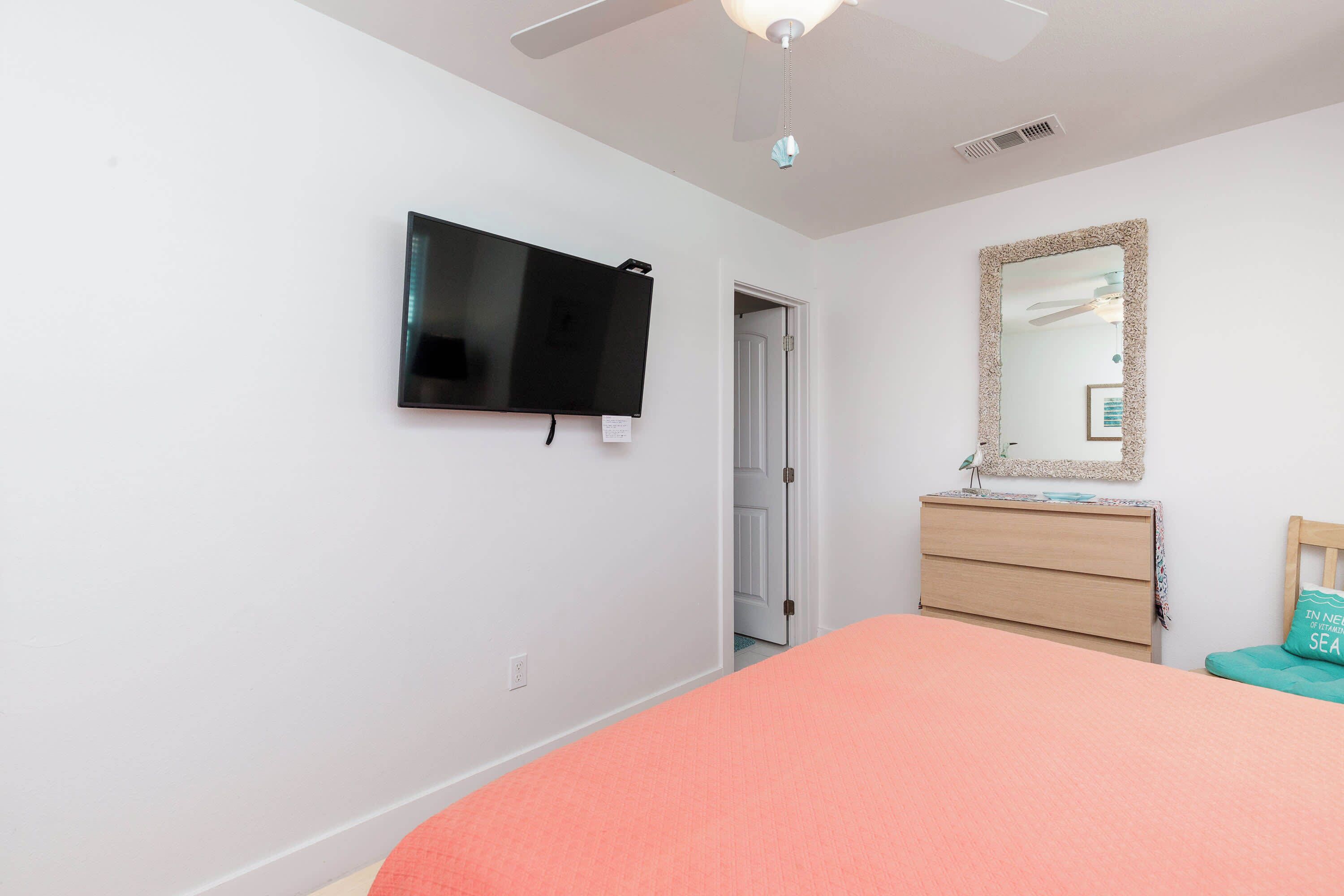 A bedroom with a bed, a television mounted on the wall, a dresser, a mirror, and a ceiling fan.
