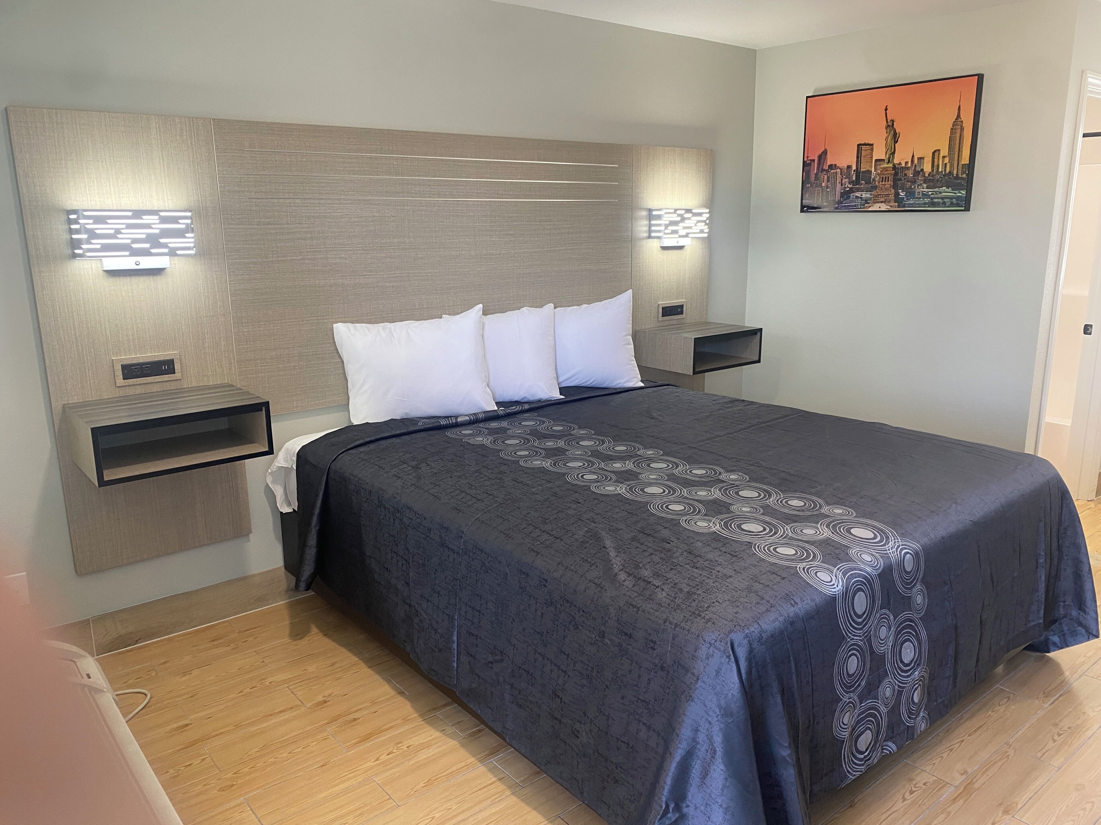 A modern hotel room with a large bed, two bedside tables, a headboard with built-in lighting, and a cityscape painting on the wall.