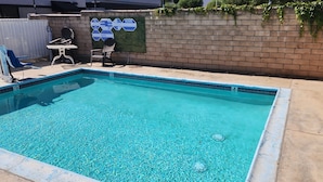 A rectangular swimming pool with clear blue water, surrounded by a concrete deck. There are lounge chairs and a table near the pool, and a brick wall with a warning sign in the background. - Mayfair Inn Ontario Airport (Ontario)