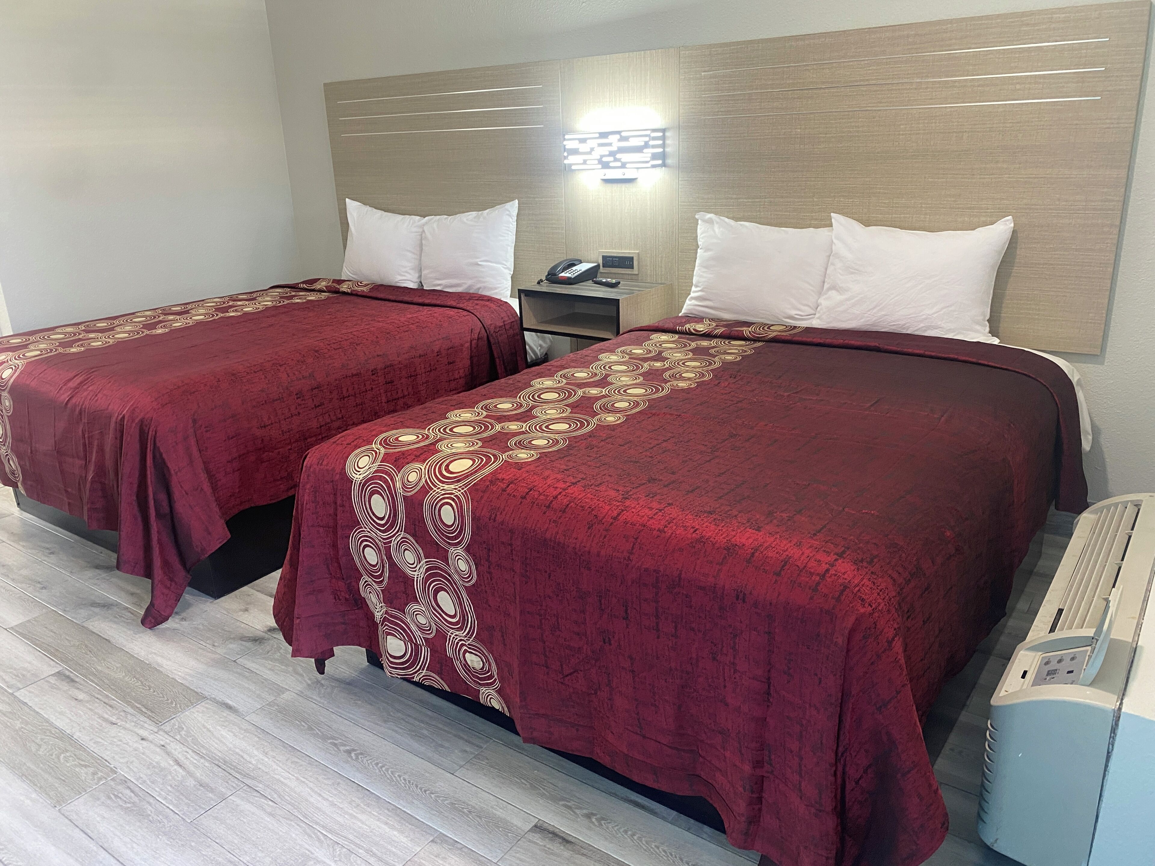 A hotel room with two beds, each with a red bedspread and white pillows. There is a bedside table with a phone and a lamp, and a wall-mounted light fixture above the beds.