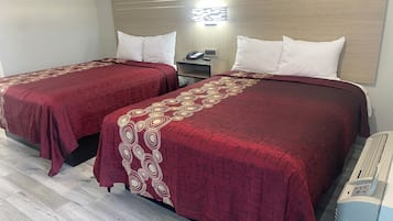 A hotel room with two beds, each with a red bedspread and white pillows. There is a bedside table with a phone and a lamp, and a wall-mounted light fixture above the beds.