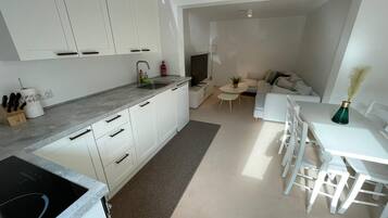 Een moderne keuken met witte kasten, een grijs aanrechtblad en een ingebouwde kookplaat. Er is een eethoek met een witte tafel en stoelen, en een klein rond tafeltje met een plant erop.