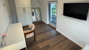 Et moderne, lyst værelse med trægulv, et hvidt skrivebord, en stol, et fjernsyn monteret på væggen og et stort vindue med udsigt til det fri.