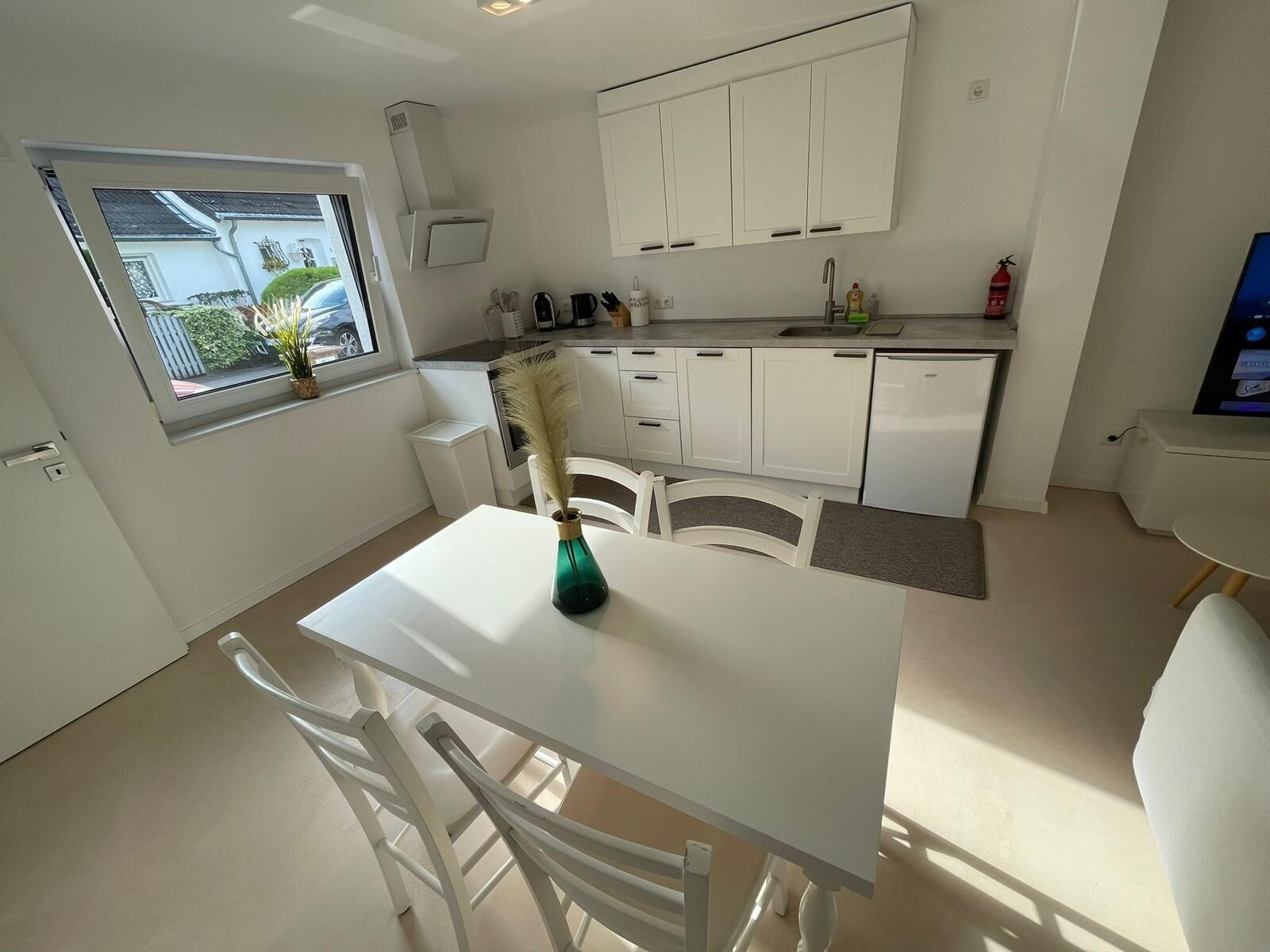 Une cuisine moderne avec des meubles blancs, une table à manger et des chaises. Une fenêtre offre une vue sur l’extérieur.