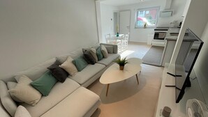 En moderne stue med sofa, sofabord og fjernsyn. I baggrunden ses et køkkenområde med et spisebord og stole.