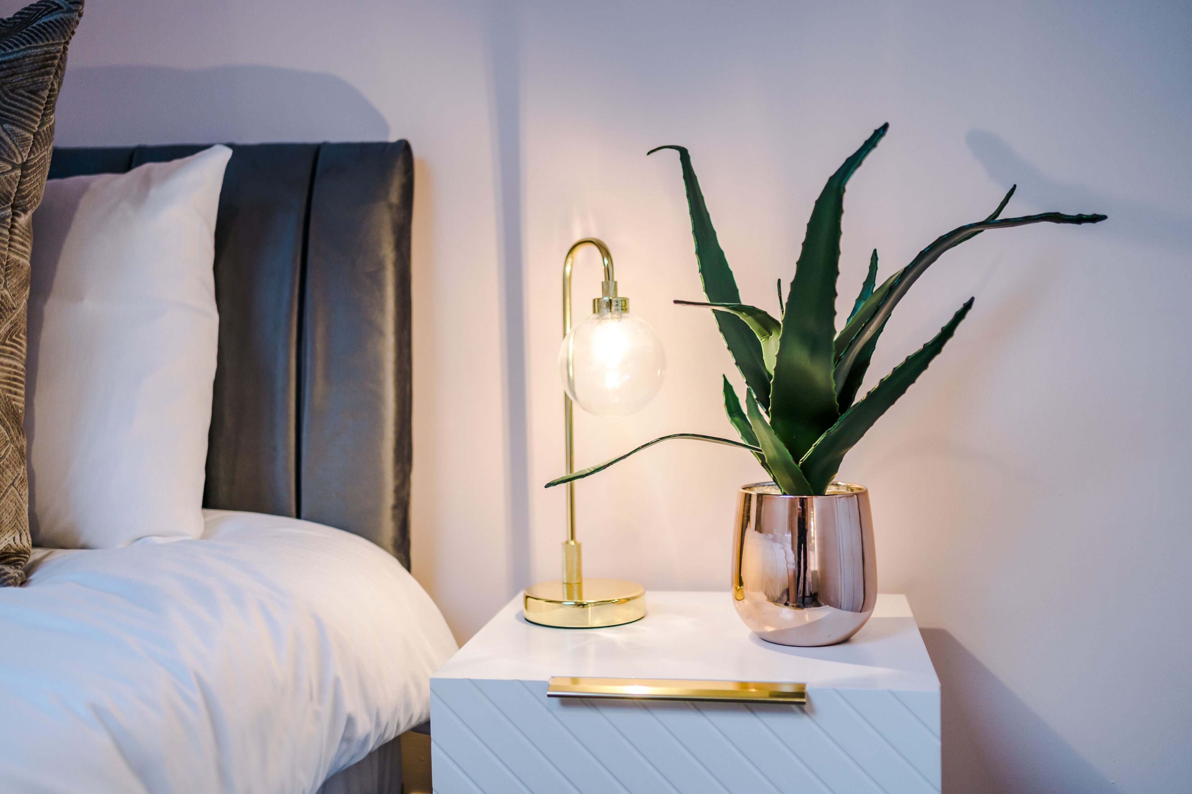A neatly made bed with white bedding, a gold lamp, and a potted plant on a white nightstand.