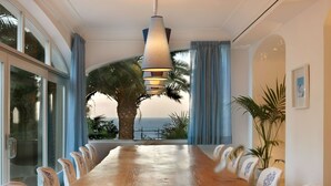 Une longue table en bois, des chaises, de grandes fenêtres et une vue sur la mer.