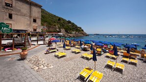 Un hôtel situé en bord de mer, doté d’une terrasse et de parasols, ainsi qu’une plage rocheuse et des bateaux amarrés dans l’eau.