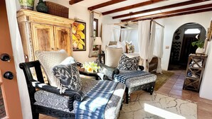 Ein geräumiges Wohnzimmer mit hohen Decken, freiliegenden Holzbalken, einem Deckenventilator und einem großen Fenster, das für natürliches Licht sorgt. Es gibt eine gemütliche Sitzecke mit gemusterten Sofas und einem dekorativen Teppich.
