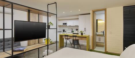 A modern hotel room with a large bed, a TV, a desk with two chairs, a kitchen area, and a bathroom.