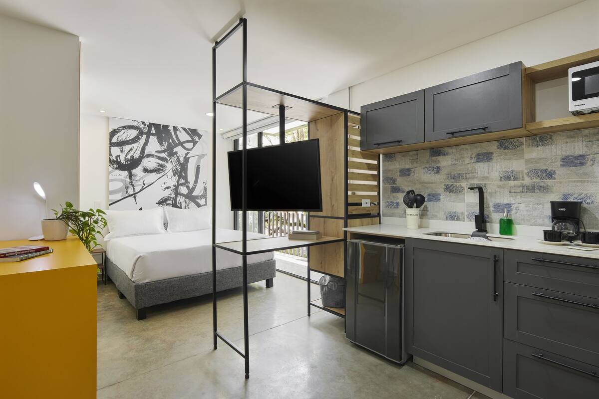 Un dormitorio moderno con cama, escritorio y un cuadro abstracto grande. La habitación tiene una kitchenette con microondas, cafetera y armarios.