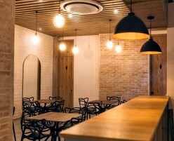 Interior de restaurante con paredes de ladrillo, vigas de madera en el techo y lámparas colgantes. Mesas y sillas dispuestas para comer.