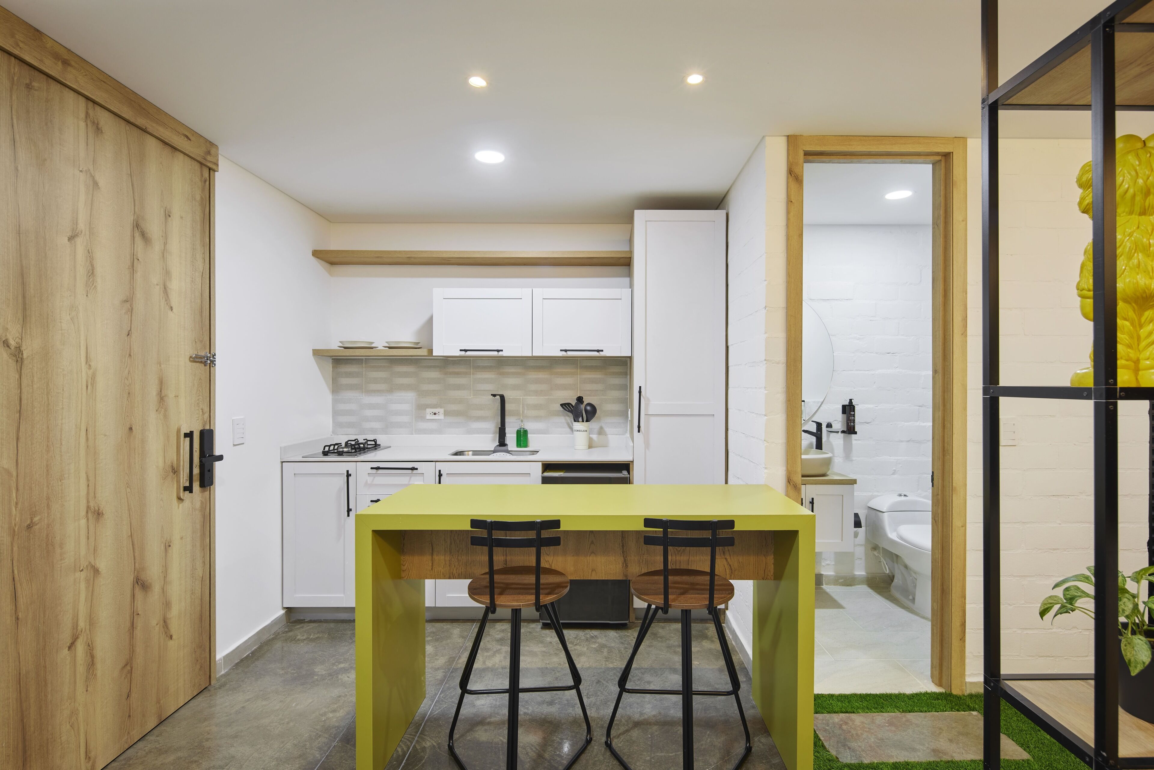 Une cuisine compacte avec des meubles blancs, un îlot jaune doté de tabourets et une porte en bois.