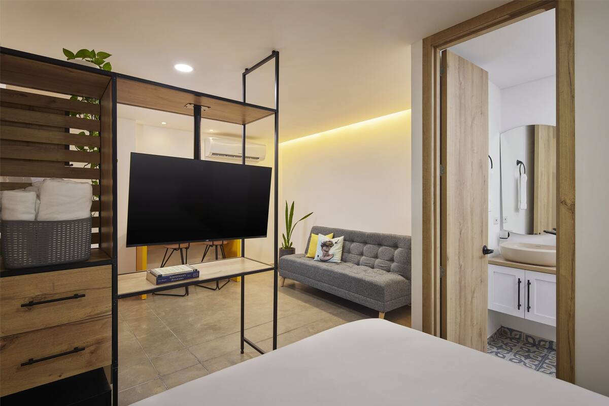 Una habitación de hotel moderna con sofá, televisor en un soporte, estante de madera con una cesta, y baño con lavamanos y espejo.