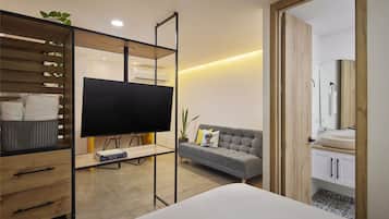 Une chambre d’hôtel moderne avec un canapé, un téléviseur posé sur un meuble, une étagère en bois avec un panier, et une salle de bain équipée d’un lavabo et d’un miroir.
