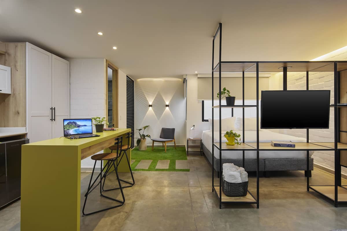 Habitación de hotel pequeña con kitchenette, un escritorio con una computadora portátil, una cama, un televisor y un área de descanso.