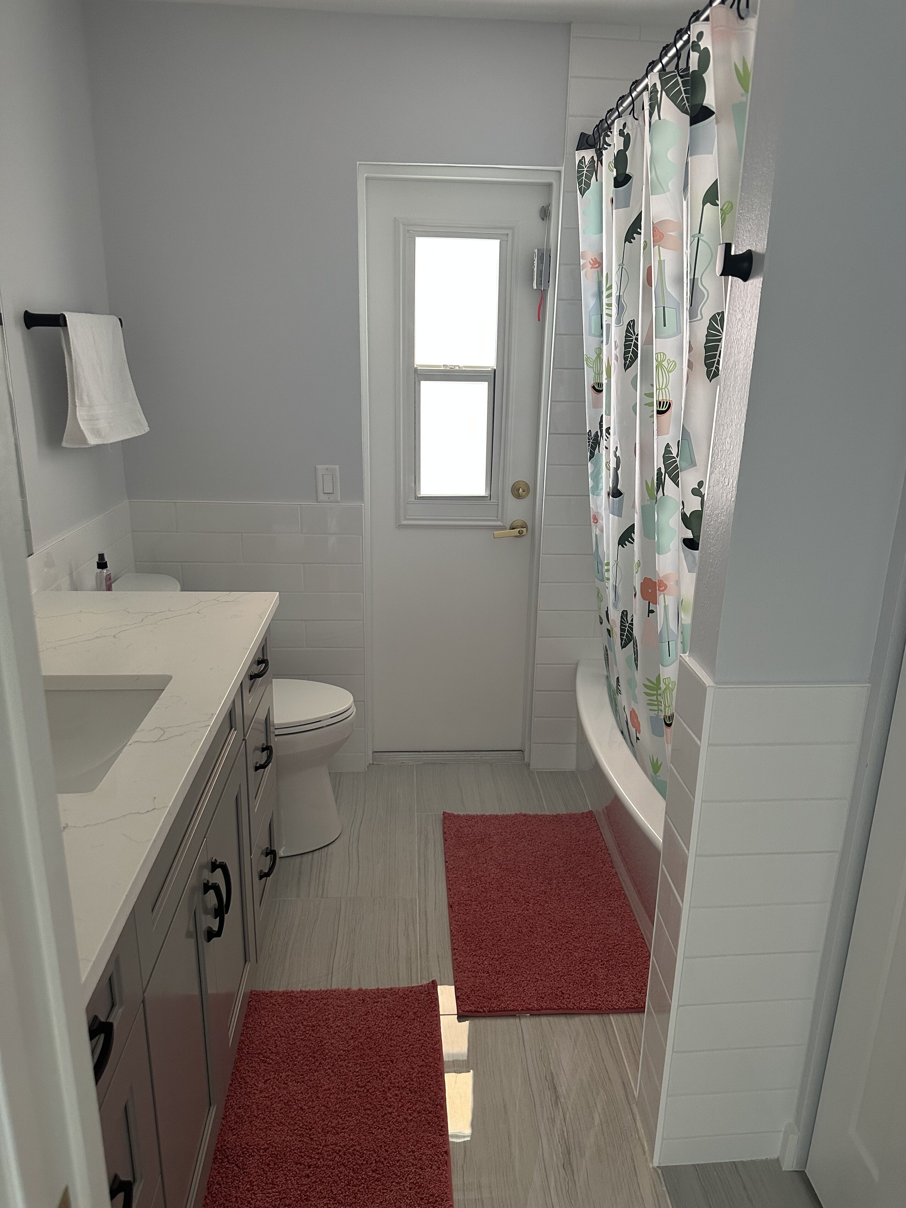 Une salle de bain avec des toilettes, un lavabo, un rideau de douche orné d’un motif végétal et deux tapis rouges.