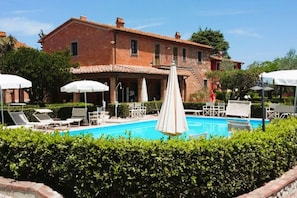 Zona de piscina con sillas de descanso y sombrillas, rodeada de setos y un edificio de ladrillo.