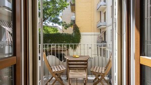 Un balcón con una mesa de madera y dos sillas, una mesita con un plato, y vistas a un edificio con balcones y vegetación.