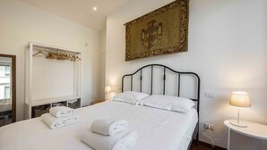 Una cama bien hecha con sábanas y toallas blancas, una mesita de noche con lámpara, un armario con ropa colgada y un tapiz en la pared.
