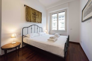 Un lit bien fait, avec du linge de lit blanc, une table de chevet avec une lampe, une fenêtre donnant sur des bâtiments et une tapisserie au mur.