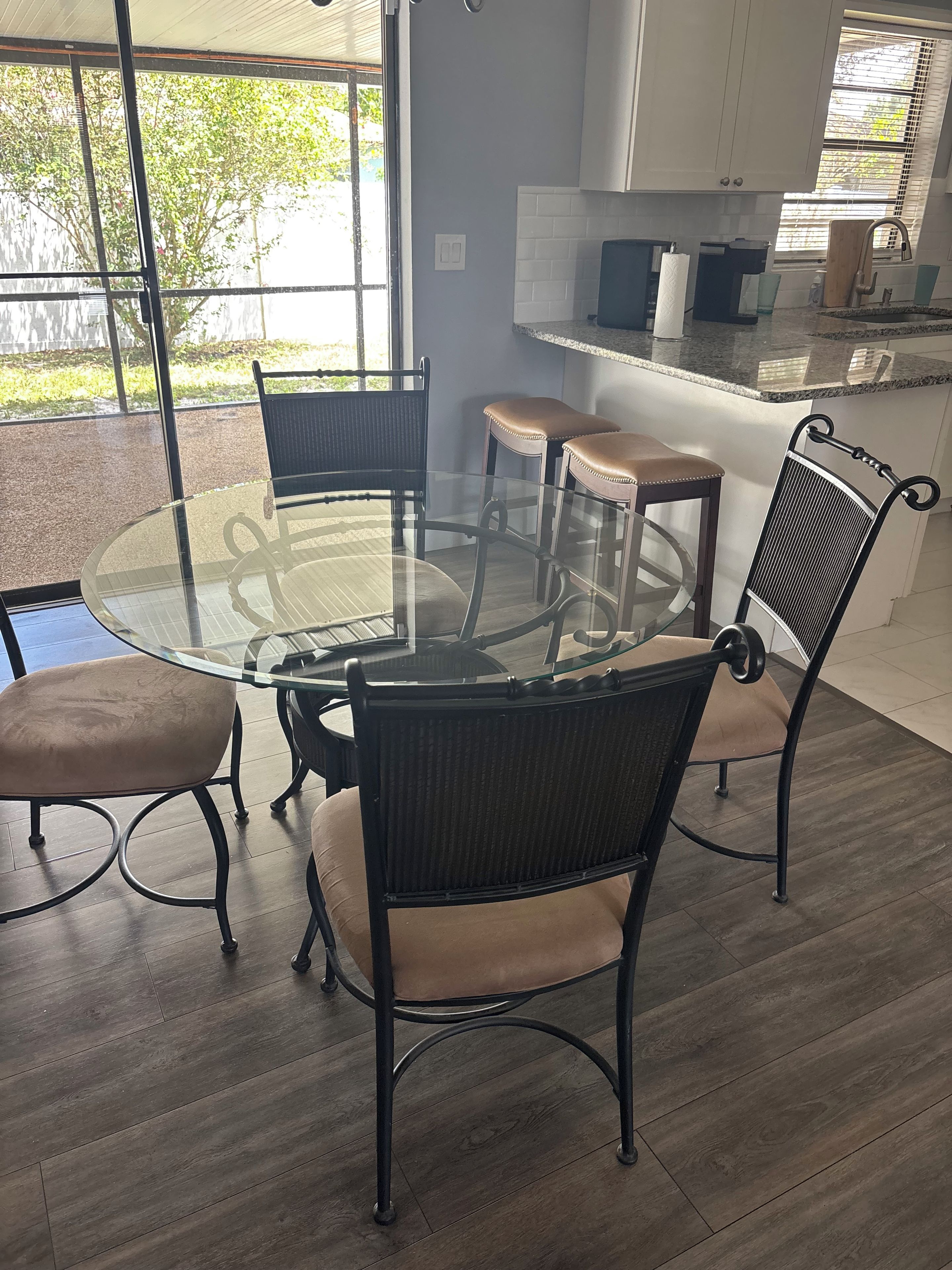 Un coin repas avec une table et des chaises à plateau en verre, une cuisine en arrière-plan et une vue sur la verdure à l’extérieur.