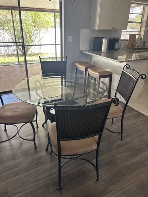 A dining area with a glass-top table and chairs, a kitchen in the background, and a view of greenery outside.
