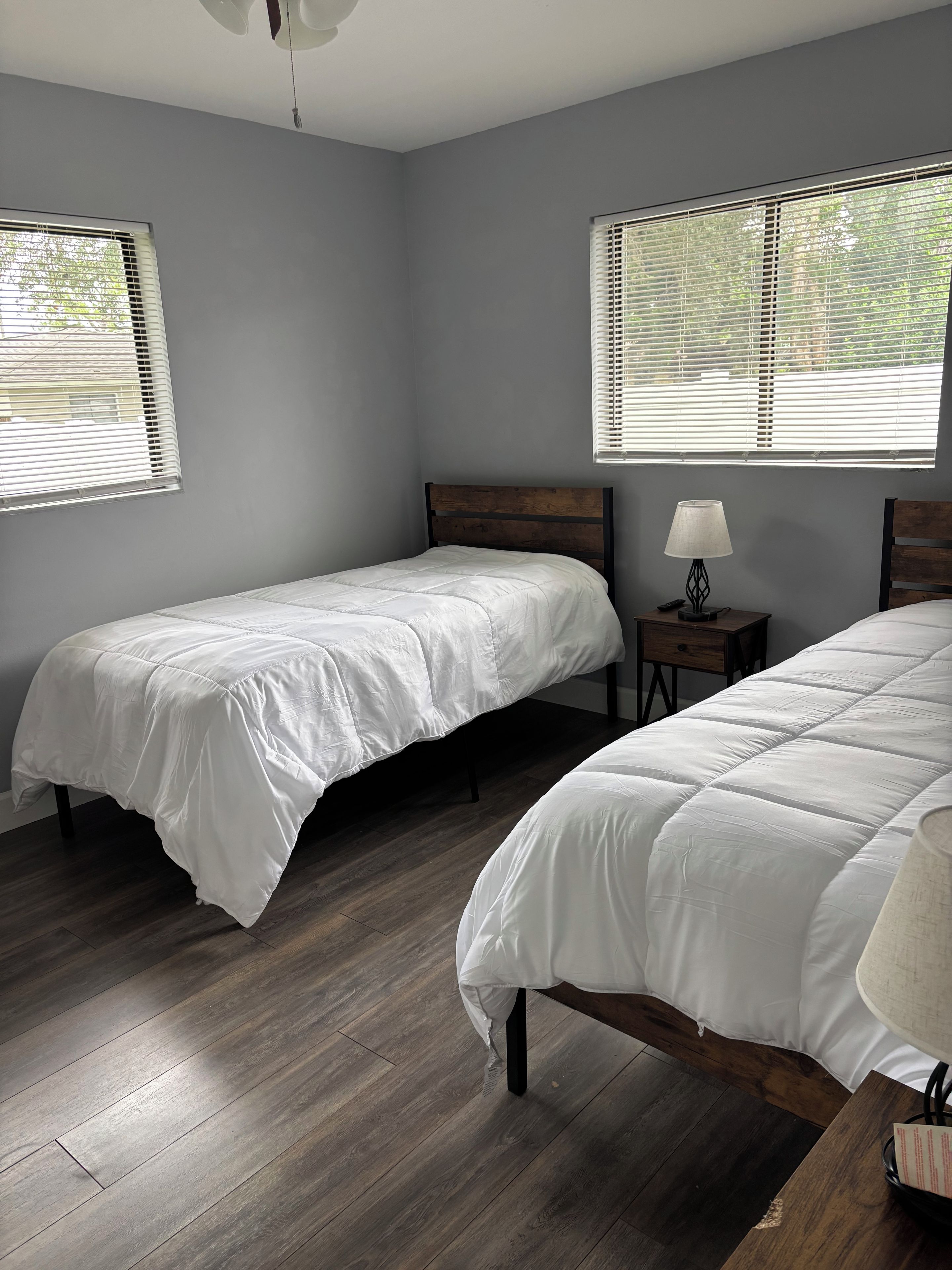 A bedroom with two single beds, a nightstand with a lamp, and a window with blinds.