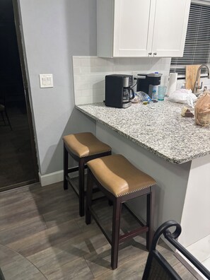 A kitchen with a granite countertop, two wooden bar stools, a coffee maker, and a trash can.