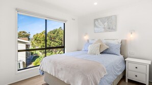 Quarto com uma grande janela, uma cama com colcha branca e azul, duas mesas de cabeceira e um aplique de parede.