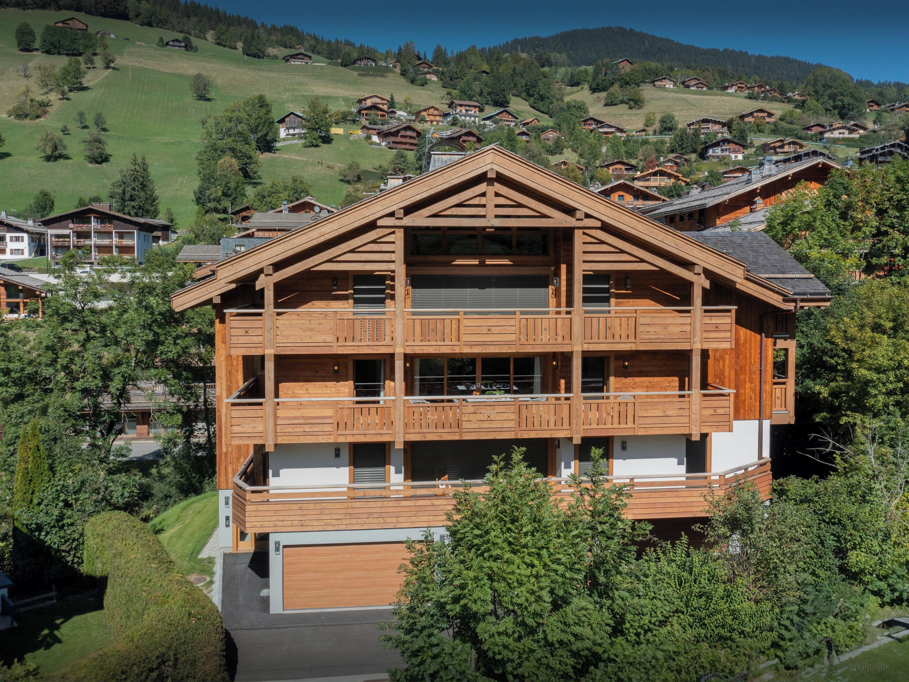 Een meerlaags houten chalet met balkon, gelegen in een bergachtig gebied met andere vergelijkbare gebouwen op de achtergrond.